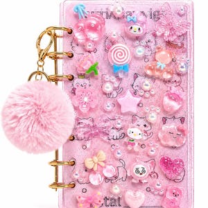 Op de afbeelding: Roze, glinsterende planner met goudkleurige ringen en een pluizige roze pompon-bedel. De planner is versierd met diverse roze bedels, waaronder hartjes, strikjes en stripfiguren. Een schattig en meisjesachtig accessoire.