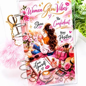 Op de afbeelding: Een doorzichtig acryl notitieboek met een kleurrijke illustratie van een persoon met lang bruin haar, omringd door make-up, bloemen en koffie. De kaft toont de tekst Women Glow Vibes, Shine, Be Confident, Stay Positive en Love Yourself. Een gouden ringband met een roze pompon sleutelhanger is bevestigd.