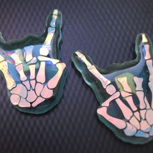 May include: Two resin-cast hand gestures, each in the shape of a rock-and-roll hand sign. The hands are translucent with a teal outline and feature a skeleton hand design in pink and white. The background is a dark, textured surface.