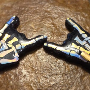 May include: Two black resin hand-shaped pendants with a skeleton bone design. The bones are white and yellow, with iridescent blue and gold accents. Each pendant has a gold-colored accent on the back of the hand.