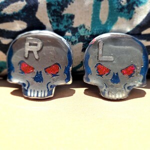 May include: Two skull-shaped decorative objects. The skulls are silver with blue accents and red glitter eyes. The letter "R" is on the left skull, and the letter "L" is on the right skull. The background is a patterned fabric.