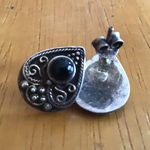 May include: A pair of silver-toned stud earrings. Each earring has a teardrop shape with a black stone center and ornate, swirling details. The earrings are displayed on a wooden surface.