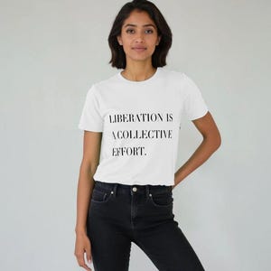 Könnte beinhalten: Ein weißes T-Shirt mit dem Text "LIBERATION IS A COLLECTIVE EFFORT" in Schwarz. Das Shirt wird mit einer schwarzen Jeans kombiniert. Der Hintergrund ist hellgrau.
