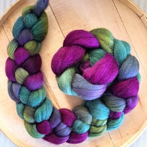 May include: A close-up of a braided fiber art piece in shades of purple, green, and teal. The fiber is arranged in a circular pattern on a wooden surface. The colors blend together, creating a visually appealing texture.