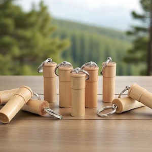 May include: A collection of small, cylindrical wooden containers with keychains. The containers are light brown and have a natural wood grain. Some are upright, while others are lying on a wooden surface. The background is a blurred view of trees.
