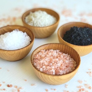 May include: Four small wooden bowls filled with different types of salt: white, pink, black, and grey.