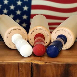 May include: Three wooden rolling pins with white, red, and blue handles are displayed. The rolling pins are made by Fletchers' Mill and Made in Maine U.S.A. The background features the American flag.