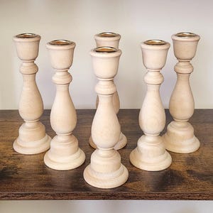 May include: Six unfinished wooden candle holders with a light wood grain. Each holder has a gold-colored metal cup at the top for holding candles. The holders have a classic, turned design with a wide base and a bulbous center.