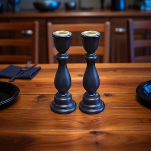 May include: Two black wooden candle holders with a classic design. The candle holders have a round base, a decorative stem, and a cup-shaped top. The tops have a gold-colored interior. The candle holders are on a wooden table.