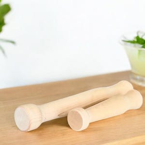 May include: Two wooden muddlers, one larger than the other, are lying on a wooden surface. The muddlers are made of light-colored wood and have a smooth, rounded surface.