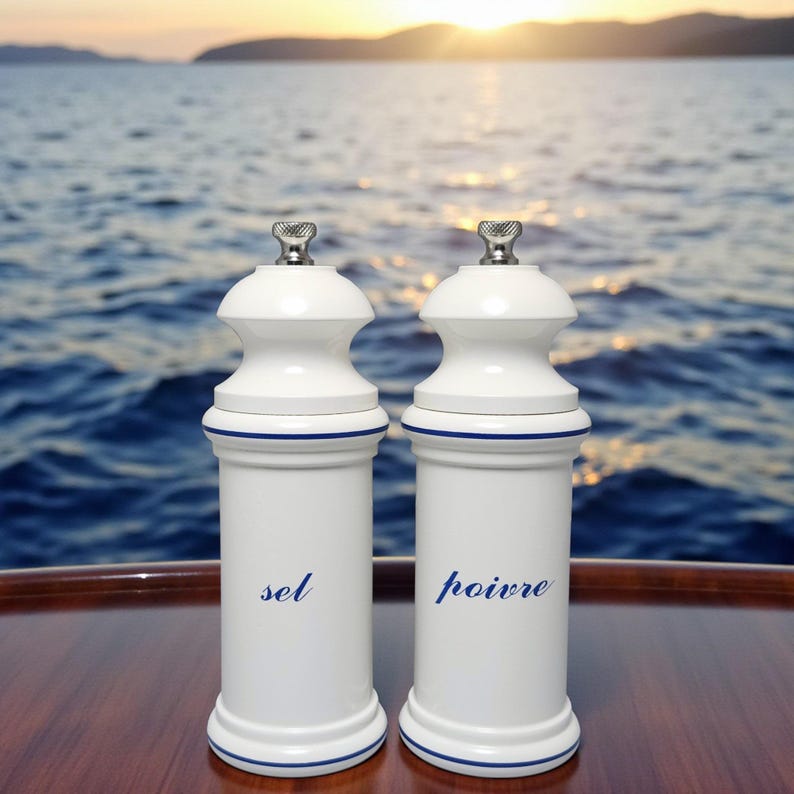 May include: Two white ceramic salt and pepper grinders with silver tops and blue trim. The word "sel" is printed on one, and "poivre" on the other. The grinders are on a wooden surface with a seascape background.
