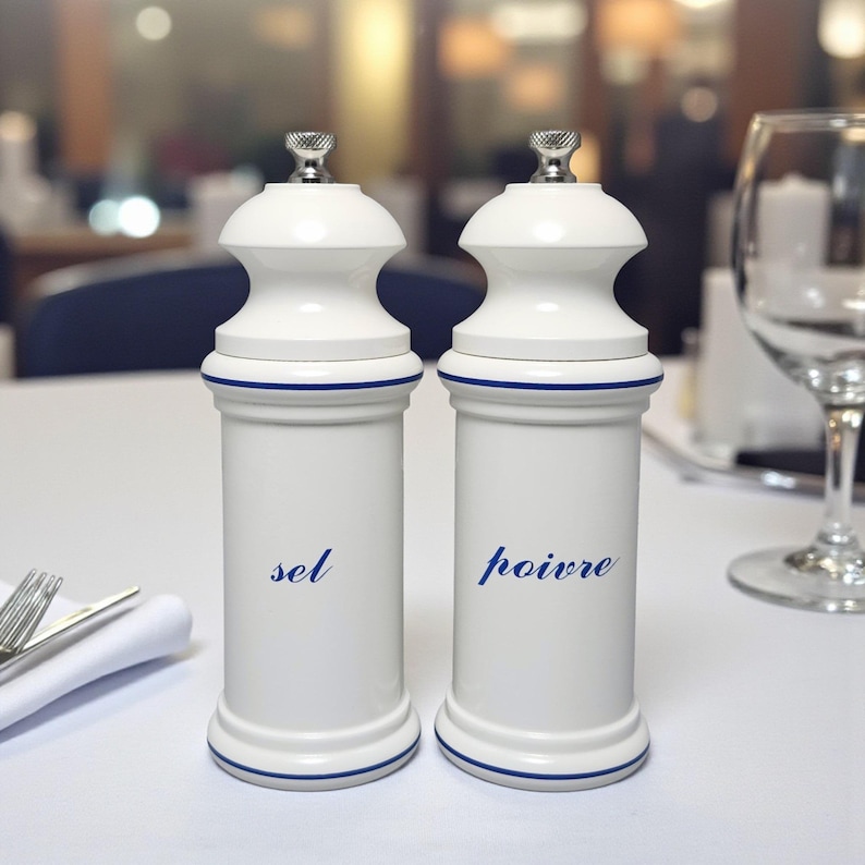 May include: Two white ceramic salt and pepper grinders with a blue accent line. The word "sel" is printed on one, and "poivre" on the other. The grinders have a silver top and are on a white tablecloth.
