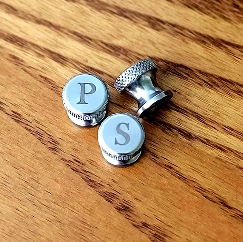 May include: Three silver-toned salt and pepper shaker tops and a decorative shaker. Two tops have the letters "P" and "S" on a light blue background. The third shaker has a decorative design. The items are on a wooden surface.