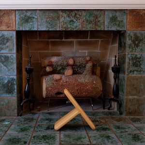 May include: A light-coloured wooden meditation stool with a curved seat and angled legs, positioned on a green tiled floor in front of a fireplace featuring a decorative tile surround and a stack of logs resting on a metal grate.