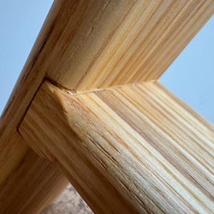 Op de afbeelding: Close-up van een houten constructie met in elkaar grijpende balken. De houtnerf is zichtbaar en toont een natuurlijke, lichtbruine kleur. De balken zijn in een hoek van 90 graden verbonden, wat het vakmanschap en de verbinding laat zien. De achtergrond is lichtblauw.