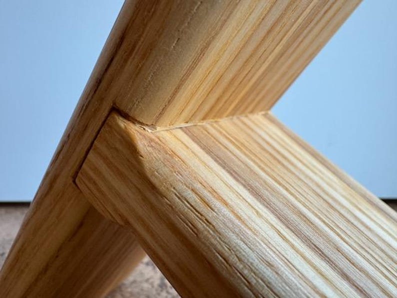 Op de afbeelding: Close-up van een houten constructie met in elkaar grijpende balken. De houtnerf is zichtbaar en toont een natuurlijke, lichtbruine kleur. De balken zijn in een hoek van 90 graden verbonden, wat het vakmanschap en de verbinding laat zien. De achtergrond is lichtblauw.
