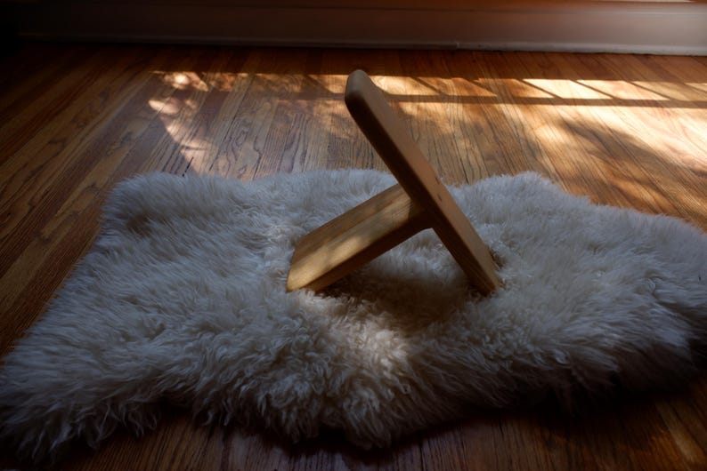 Op de afbeelding: Een houten standaard met een T-vormig ontwerp ligt op een witte, zachte vloerkleed. De standaard is gemaakt van lichtgekleurd hout en is iets schuin geplaatst. Het vloerkleed ligt op een houten vloer, met zonlicht dat de sc&egrave;ne overspoelt.