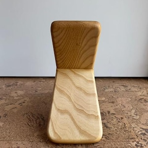 Op de afbeelding: Een lichtgekleurde houten stoel met een gebogen rugleuning en een brede, schuine zitting. De stoel is gemaakt van lichtgekleurd hout met zichtbare nerfpatronen. De stoel is ontworpen met een minimalistische esthetiek.