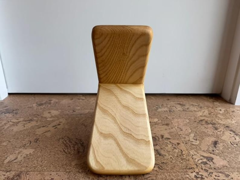Op de afbeelding: Een lichtgekleurde houten stoel met een gebogen rugleuning en een brede, schuine zitting. De stoel is gemaakt van lichtgekleurd hout met zichtbare nerfpatronen. De stoel is ontworpen met een minimalistische esthetiek.