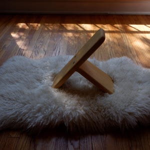 Op de afbeelding: Een houten boekenstandaard in X-vorm ligt op een witte, donzige schapenvacht. De vacht ligt op een houten vloer, met zonlicht dat schaduwen werpt. De standaard is gemaakt van lichtgekleurd hout.