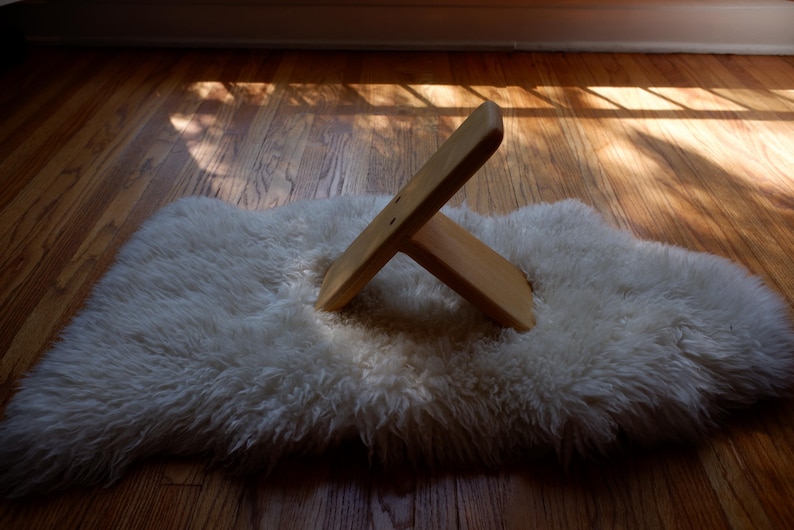 Op de afbeelding: Een houten boekenstandaard in X-vorm ligt op een witte, donzige schapenvacht. De vacht ligt op een houten vloer, met zonlicht dat schaduwen werpt. De standaard is gemaakt van lichtgekleurd hout.