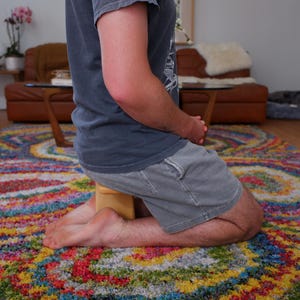 Op de afbeelding: Een persoon knielt op een kleurrijk tapijt met een houten blok onder de knie&euml;n. Het tapijt heeft een levendig patroon van rood, geel, blauw en groen. De persoon draagt een grijs t-shirt en een korte broek.