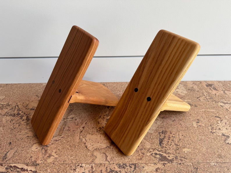 May include: Two angled wooden stands with a natural light brown wood grain finish. Each stand features a rectangular front panel with two small, dark circular holes positioned horizontally near the center, supported by a triangular base piece.