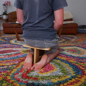 Op de afbeelding: Een persoon knielt op een kleurrijk, patroonrijk tapijt en gebruikt een kleine houten kruk. Het tapijt heeft een levendig wervelontwerp met meerdere kleuren. De persoon draagt een grijs t-shirt en een korte broek. Op de achtergrond staat een bruine leren bank.