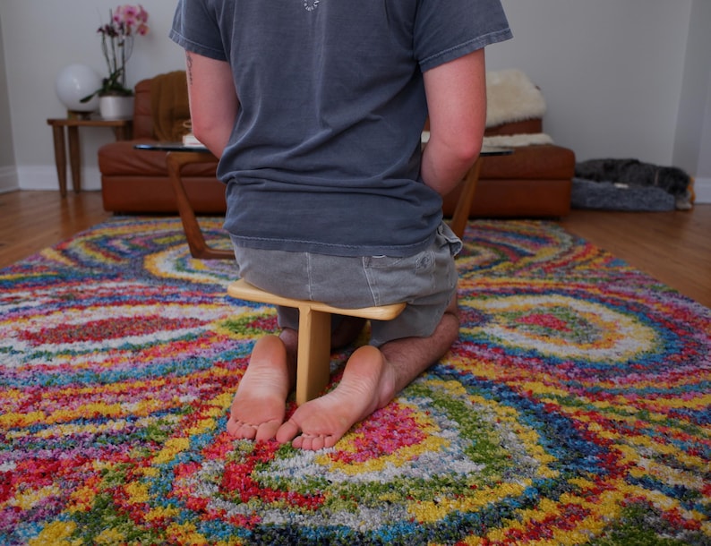Op de afbeelding: Een persoon knielt op een kleurrijk, patroonrijk tapijt en gebruikt een kleine houten kruk. Het tapijt heeft een levendig wervelontwerp met meerdere kleuren. De persoon draagt een grijs t-shirt en een korte broek. Op de achtergrond staat een bruine leren bank.