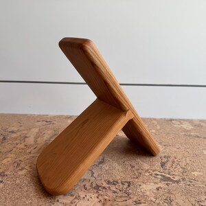 May include: A light brown wooden stand featuring two interlocking rectangular pieces. The design consists of a vertical back panel and a shorter, angled front support piece, both displaying a smooth, natural wood grain finish throughout.