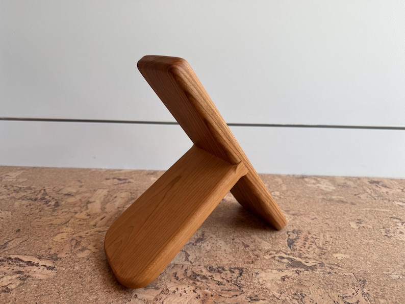 May include: A light brown wooden stand featuring two interlocking rectangular pieces. The design consists of a vertical back panel and a shorter, angled front support piece, both displaying a smooth, natural wood grain finish throughout.