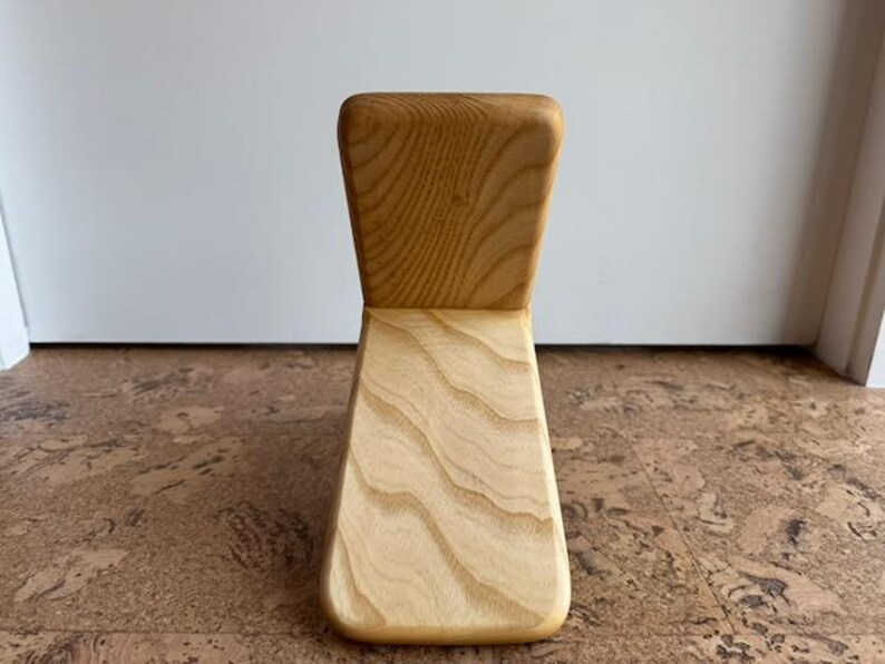 May include: A light-colored wooden meditation chair featuring a smooth, curved seat and a vertical backrest. The natural wood grain is clearly visible across the entire surface, showcasing a light tan hue with darker, flowing patterns throughout.