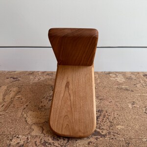 May include: A small, L-shaped wooden object crafted from light-toned wood with a visible grain pattern. The piece features a vertical backrest section and a flat, horizontal base section, both joined together to form a singular, smooth, and solid structure.