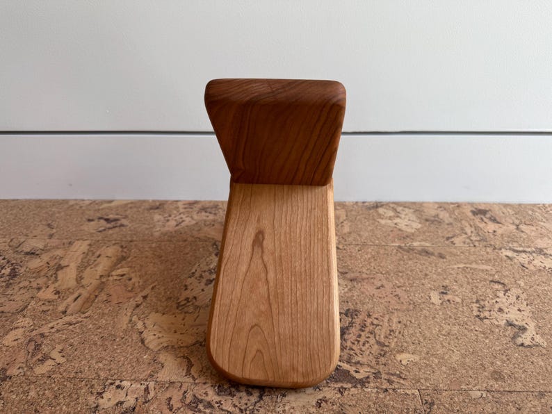 May include: A small, L-shaped wooden object crafted from light-toned wood with a visible grain pattern. The piece features a vertical backrest section and a flat, horizontal base section, both joined together to form a singular, smooth, and solid structure.