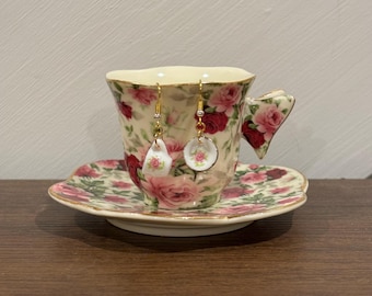 May include: A floral teacup, saucer, and matching earrings. The teacup and saucer are decorated with pink roses and green leaves. The earrings have a teardrop and circular design with a floral pattern, suspended from gold-coloured hooks.