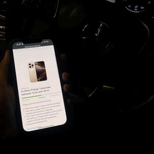 May include: A smartphone screen displaying a confirmation message for an in-store pickup order for a gold iPhone. The message includes the pickup date, time, and instructions for the pickup process. The phone is held in a hand inside a car.
