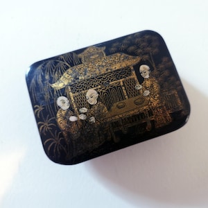 Könnte beinhalten: Rechteckige schwarze Box mit einem gold-schwarzen asiatisch inspirierten Design. Die Box zeigt eine detaillierte Szene mit Figuren und einer Pagodenstruktur. Das Kunstwerk ist aufwendig und die Box hat eine glänzende Oberfläche.
