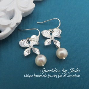 May include: Silver flower earrings with a pearl drop. The earrings are on a blue background.