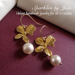 May include: Gold-toned flower earrings with a dangling white pearl. The earrings have a hook closure and are displayed on a purple surface. Text on the image reads "Sparklies by Julie Unique handmade jewelry for all occasions."