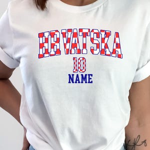 May include: A white short-sleeved t-shirt featuring the text HRVATSKA in a red and white checkered pattern, with the number 10 and the word NAME printed in blue below it. The shirt is tied at the waist and paired with light blue denim jeans.