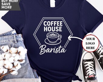 May include: Navy blue t-shirt with a white graphic. The graphic features a coffee cup with latte art inside a hexagon with the words "COFFEE HOUSE" and "Barista". The shirt is paired with denim shorts and black sneakers.