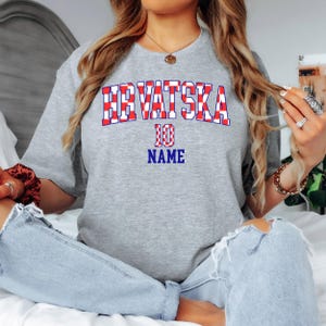 May include: A heather gray short-sleeve t-shirt featuring the text HRVATSKA in a red and white checkered pattern, with the number 10 and the word NAME printed in blue below the text in a bold, athletic-style font on the front of the shirt.