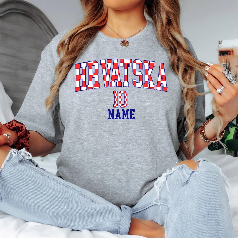May include: A heather gray short-sleeve t-shirt featuring the text HRVATSKA in a red and white checkered pattern, with the number 10 and the word NAME printed in blue below the text in a bold, athletic-style font on the front of the shirt.
