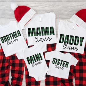 May include: A collection of white t-shirts and a baby bodysuit featuring green and black plaid text that reads Brother Claus, Mama Claus, Daddy Claus, Mini Claus, and Sister Claus, accompanied by two red and white knit winter hats.