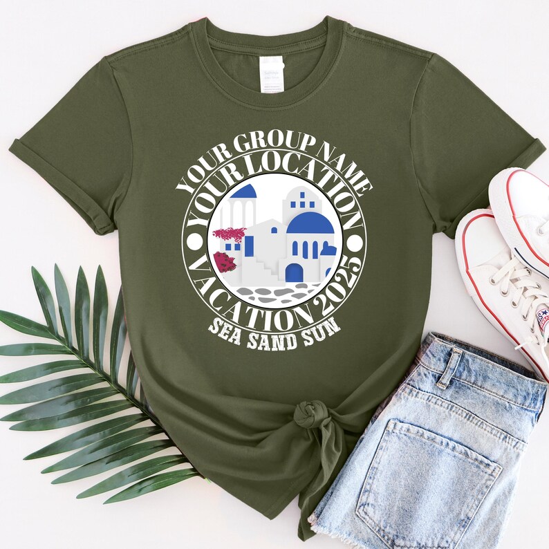 May include: An olive green t-shirt with a circular graphic. The graphic includes the text "YOUR GROUP NAME YOUR LOCATION VACATION 2025 SEA SAND SUN" and a design of white buildings with blue domes. White trainers and denim shorts are also visible.