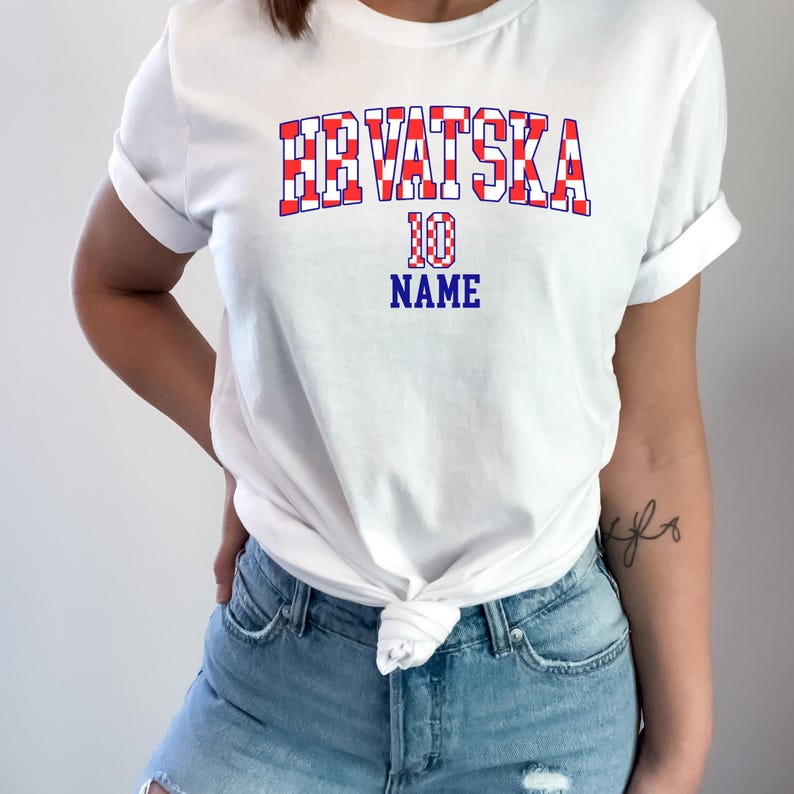May include: A white short-sleeved t-shirt featuring the text HRVATSKA in a red and white checkered pattern, with the number 10 and the word NAME printed in blue below it. The shirt is tied at the waist and paired with light blue denim jeans.