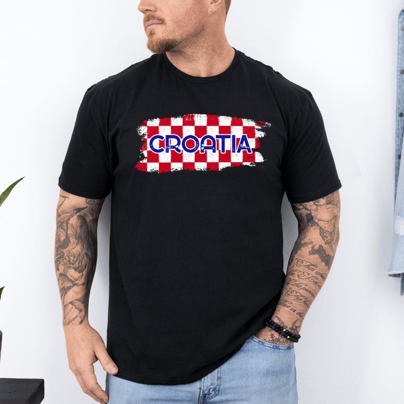 May include: A black short-sleeved t-shirt featuring a central graphic of a red and white checkered pattern with a distressed, brushstroke-style border. The word CROATIA is printed in bold, blue, outlined capital letters across the center.