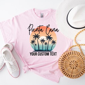 Op de afbeelding: Een lichtroze t-shirt met een afbeelding van een zonsondergang boven de oceaan met silhouetten van palmbomen. De tekst "Punta Cana" staat in zwarte schrijfletters erboven en "YOUR CUSTOM TEXT" in zwarte blokletters eronder.