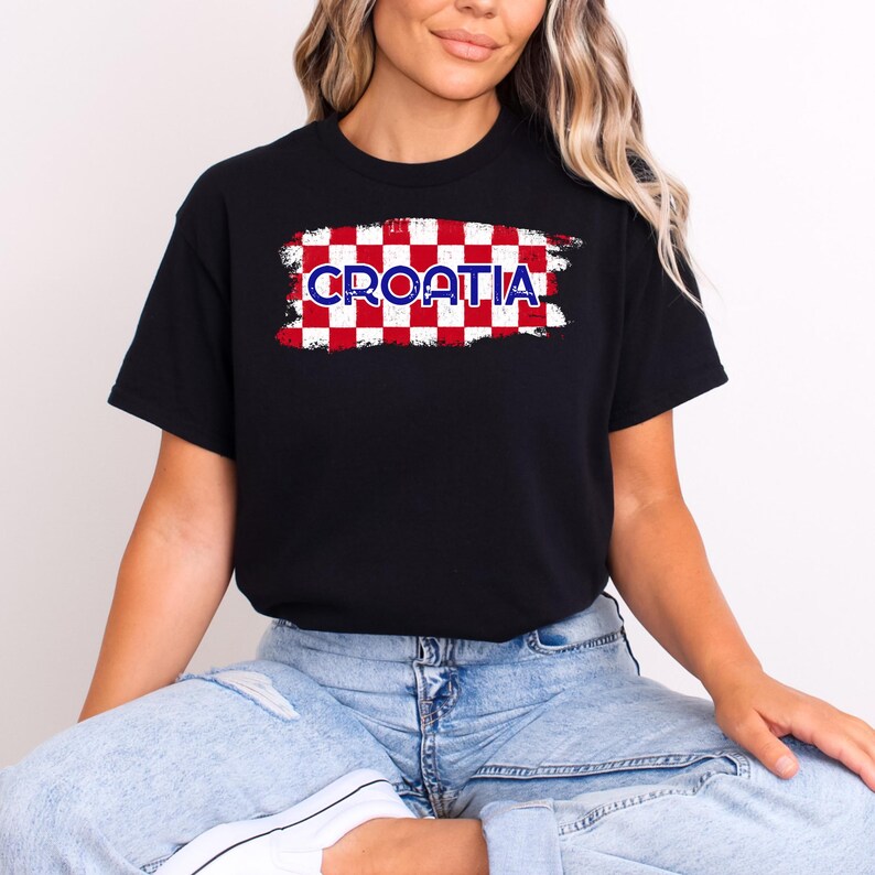 May include: A black short-sleeved t-shirt featuring a central graphic of a red and white checkered pattern with a distressed brushstroke effect. The word CROATIA is printed in blue capital letters across the center of the checkered design.