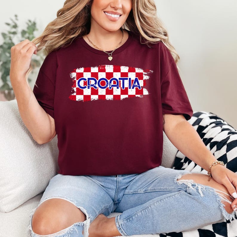 May include: A maroon short-sleeved t-shirt featuring a central graphic of a red and white checkered pattern with a distressed brushstroke effect. The word CROATIA is printed in bold blue letters with a white outline across the center of the design.
